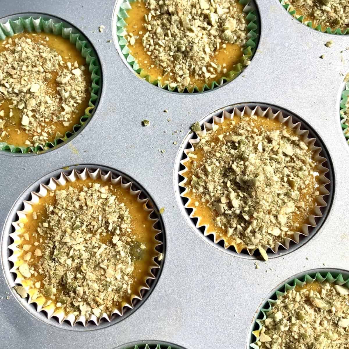 A muffin tin filled with pumpkin muffin batter topped with pumpkin seed crunch topping and ready to bake.
