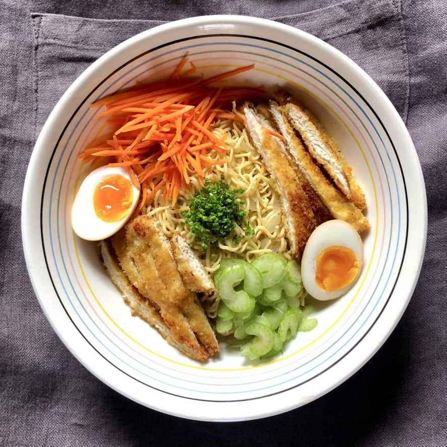 a ramen bowl with a pile of just cooked egg noodles (that look like ramen noodles) in the center with sliced chicken cutlets on opposite sides of the bowl and carrots and celery on the other opposite sides and two halves of ramen eggs added with chives in the middles