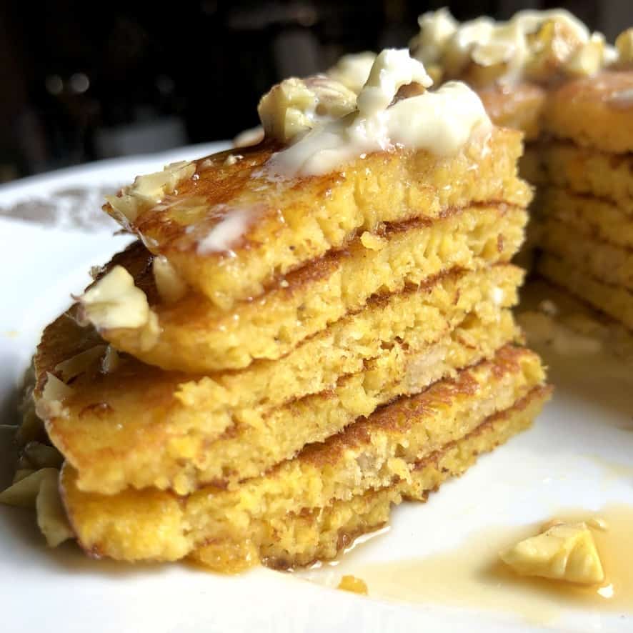 a serving platter with a stack of beautiful pumpkin-colored pancakes with melting butter, toasted chestnuts and covered with maple syrup and a wedge cut out to see the fluffy layers
