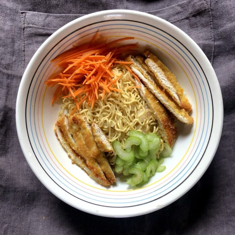 a ramen bowl with a pile of just cooked egg noodles (that look like ramen noodles) in the center with sliced chicken cutlets on opposite sides of the bowl and carrots and celery on the other opposite sides