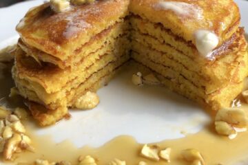 a serving platter with a hot stack of pumpkin pancakes with melting butter, toasted chestnuts and covered with maple syrup and a wedge cut out to see the fluffy layers.