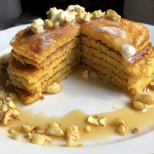 a serving platter with a hot stack of pumpkin pancakes with melting butter, toasted chestnuts and covered with maple syrup and a wedge cut out to see the fluffy layers.