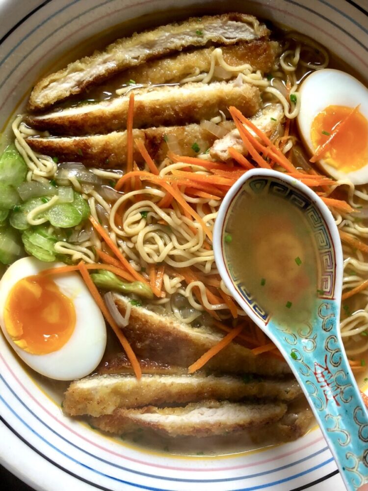 homemade chicken noodle oup with homemade noodles, sliced breaded chicken cutlet , two halves of ramen eggs with soft jammy yolks, julenned carrots, paper-thin slices of celery, and homemade swimming in homemade chicken stock with a ramen spoon