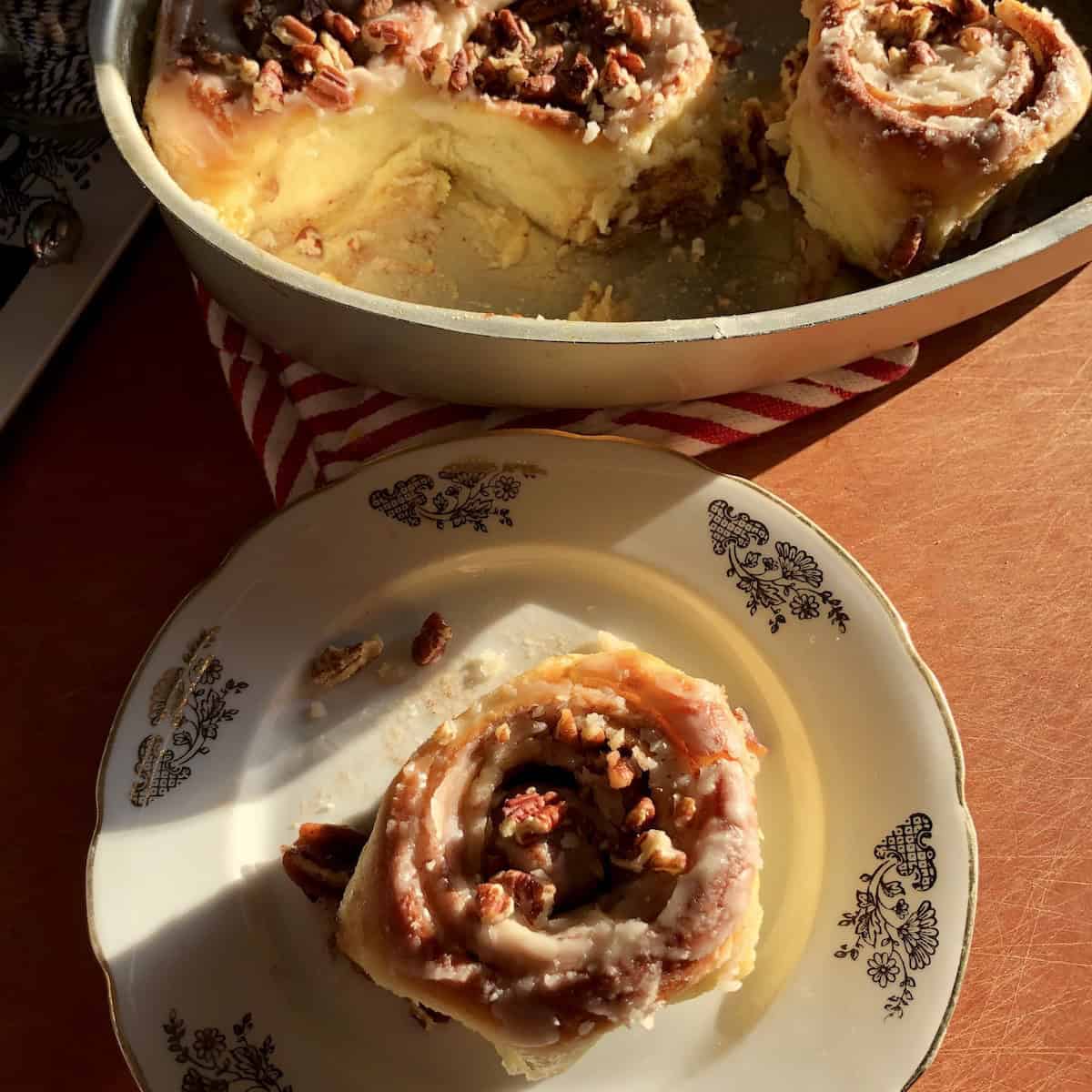 A single fluffy cinnamon roll on a dessert plate with the pan of cinnamon rolls visible in the background.
