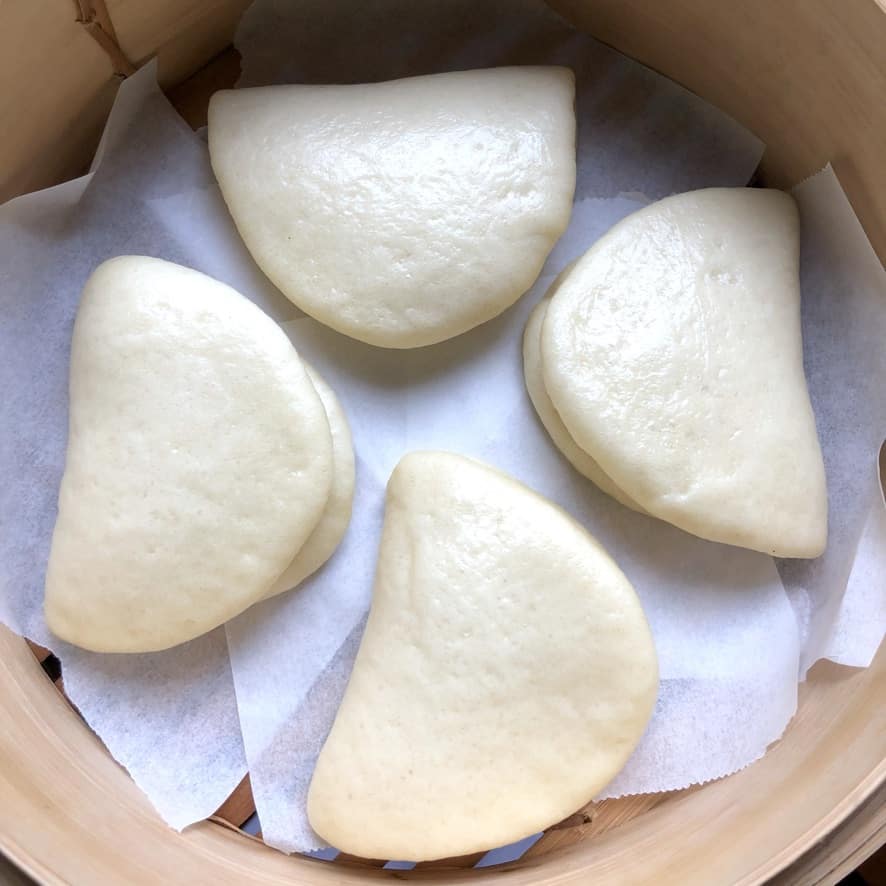 4 homemade white half-moon shaped steamed buns for pork buns in a steamer basket after just being reheated