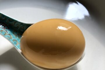 closeup of a super shiny single soft boiled caramel colored ramen egg resting in a ramen spoon in an empty ramen bowl