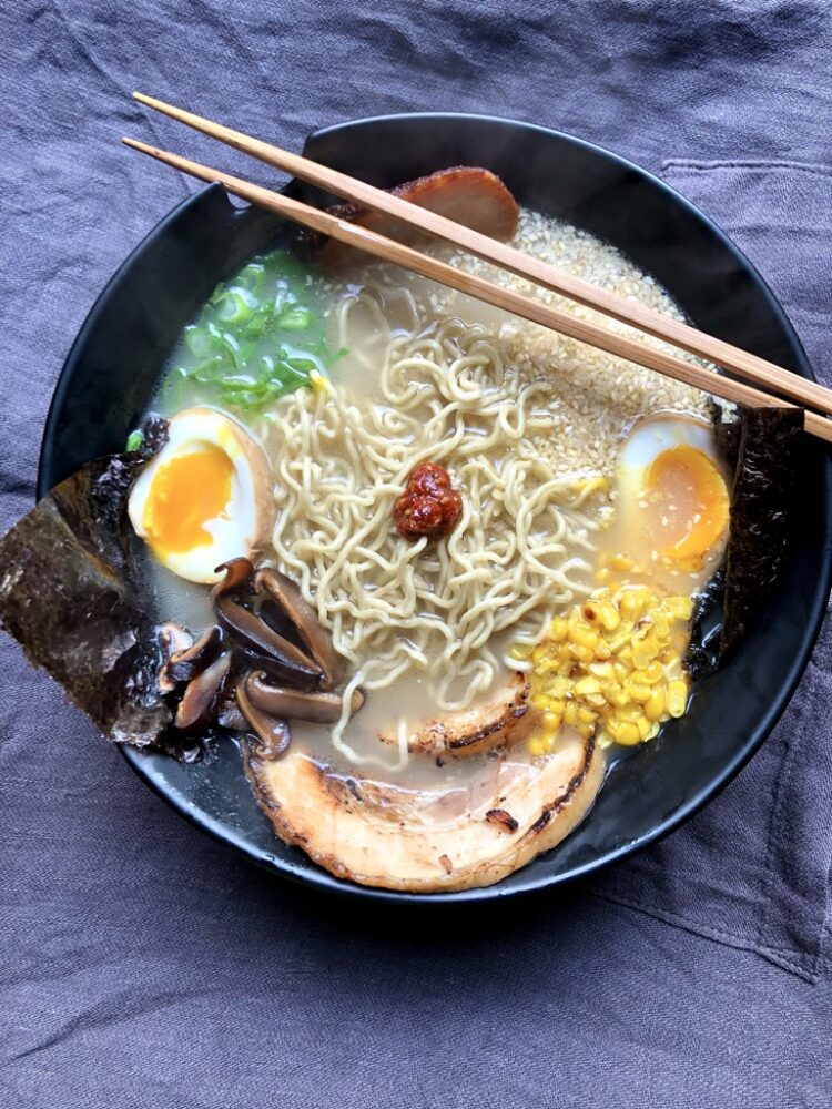 a piping hot bowl of tonkostu ramen with homemade ramen noodles, chashu pork, pork bellly, seared corn, scallions, braised pork belly, seaweed, sliced chashu braised shitake musthrooms, toasted sesame seeds, sliced scallions and