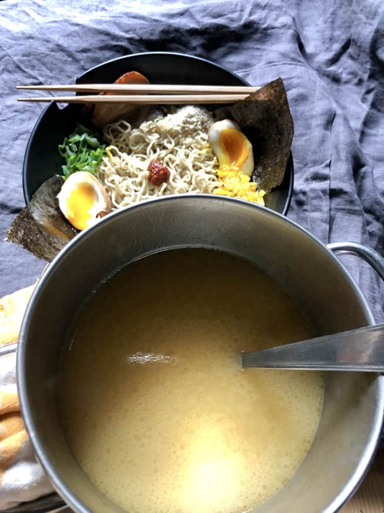 a bowl of ramen toppings with a homemade tonkotsu broth next to it about to be ladled into the bowl.