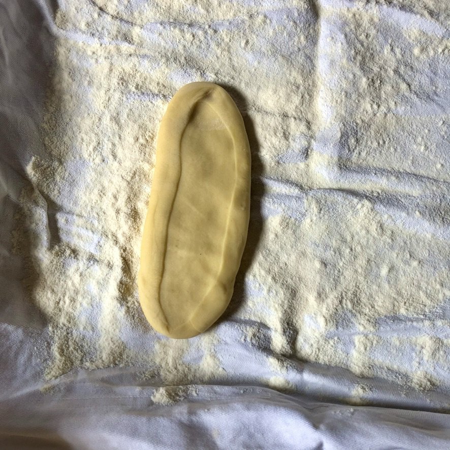 a dough piece pressed down into a long rectangular piece to make it easier to put through the pasta machine to roll it -- getting a coating of semolina before being rolled so it doesn't stick to the pasta machine