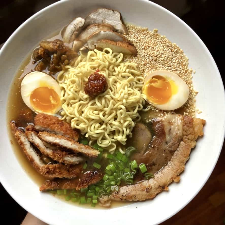 a beautiful bowl of homemade Japanese Ramen with jammy sory-mirin soft boiled ramen eggs, sliced porchetta, sliced fresh shitake mushrooms, yellow squiggly ramen noodles, roast pork belly, chives, ramen seasoning, toasted sesame seeds, sliced panko-crusted turkey cutlet, a dab of hot sauce in the middle all in an 8 hour pork bone ramen broth