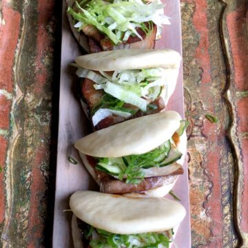 a tray with four pork buns each with two pieces of braised pork belly, quick salt and sugar pickled cumbers, sesame-soy-mayo, hoisin sauce, and scallions topped with a little shredded lettuce
