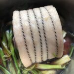 rolled pork belly tied with black and white butcher's twine resting in a pot full of braising liquid with aromatics (skin side view)