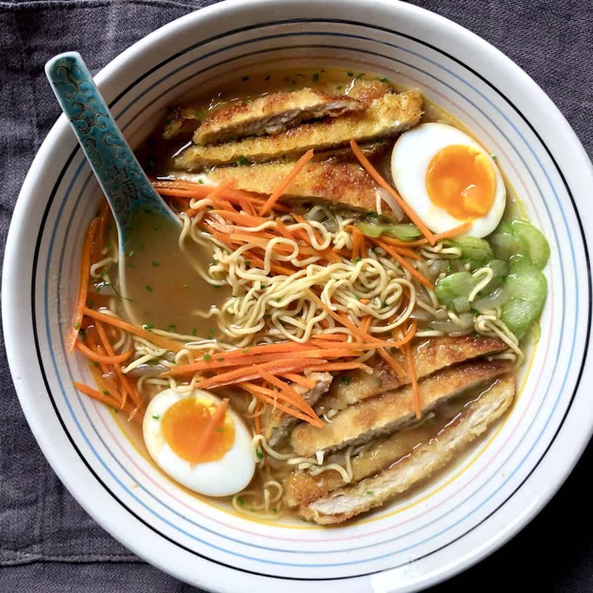 Bowl of homemade chicken noodle soup with ramen noodles, chicken katsu and ramen eggs plus sliced celery, julienned carrots, and toasted sesame seed for extra crunch.