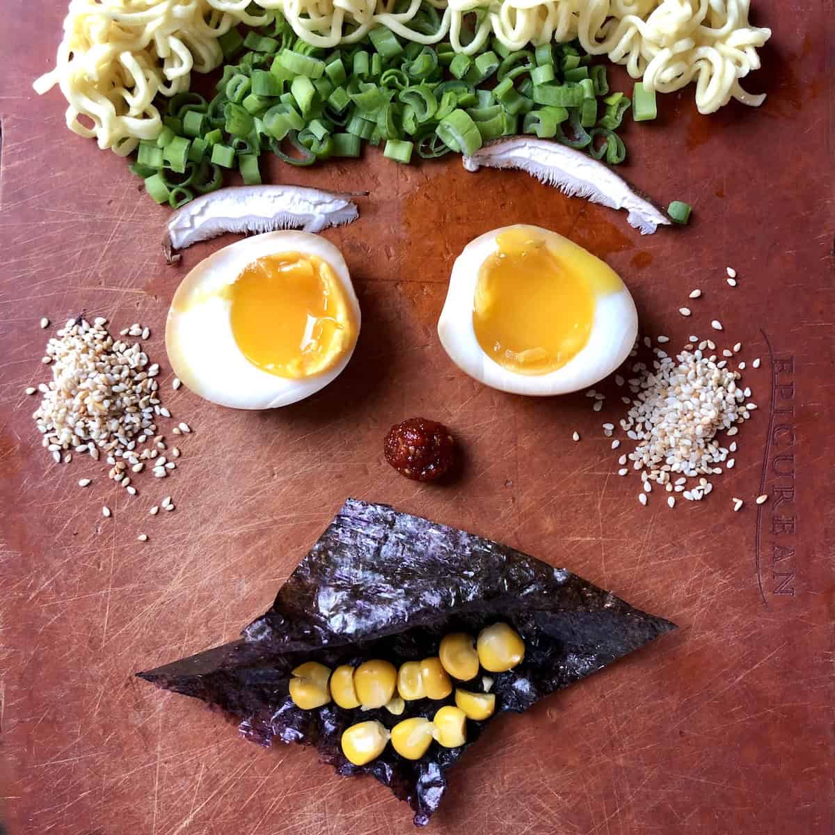Authentic ramen toppings displayed on a cutting board to look like a face with curly hair ramen noodles, scallions headband, shiitake mushroom eyebrows, ramen egg eyes, sesame seed ears, Shichimi Togarashi chili paste nose, nori mouth with corn teeth.