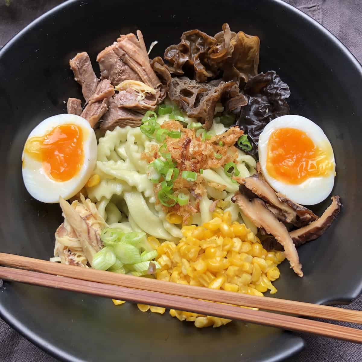 Bowl of homemade spinach udon noodles with ramen toppings ready for broth including roasted chicken roasted pork ramen eggs woodear mushrooms shiitake mushrooms scallions and corn showing assembled ramen bowl before adding chicken broth (homemade udon noodles recipe) showing ramen toppings arrangement.