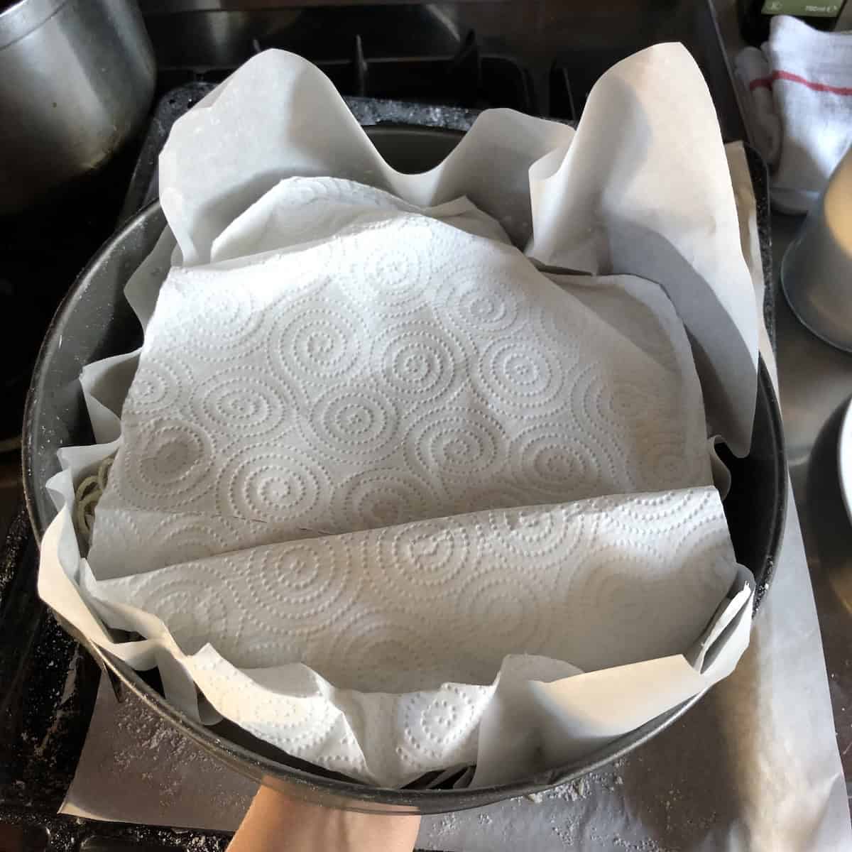 Storing finished ramen noodle nests in parchment paper lined springform pan with cornstarch dusting and paper towels on top to absorb moisture before covering with cling film and refrigerating overnight for best flavor and best method for storing homemade ramen noodles.
