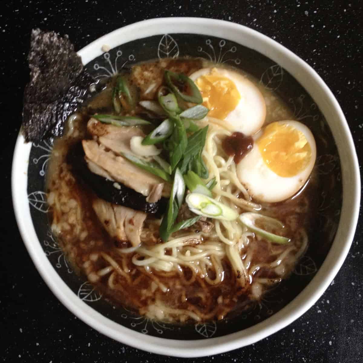 Japanese soy sauce egg halves swimming in a bowl of homemade 8-hour tonkotsu ramen broth with homemade ramen toppings including braised pork belly, chashu pork slices, scallions, sesame seeds.
