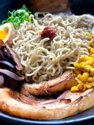 Homemade ramen noodles in a ramen bowl assembled with all toppings but no broth showing homemade ramen noodles ramen eggs chashu pork braised pork belly chives seaweed corn sesame seeds shiitake mushrooms spicy sauce ready for tonkotsu broth homemade ramen recipe complete toppings arrangement.