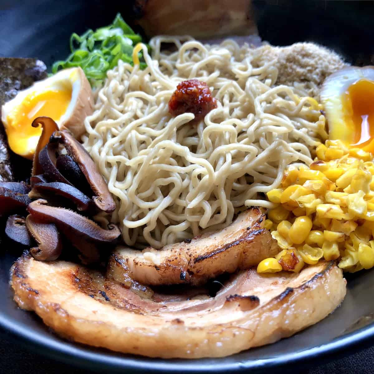 Homemade ramen noodles in a ramen bowl assembled with all toppings but no broth showing homemade ramen noodles ramen eggs chashu pork braised pork belly chives seaweed corn sesame seeds shiitake mushrooms spicy sauce ready for tonkotsu broth homemade ramen recipe complete toppings arrangement.
