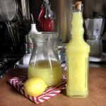 a pitcher half full milky-looking bright yellow homemade Italian limoncello next to a bottle of homemade limoncello and a fresh lemon on a red and white striped pot holder