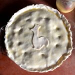 An assembled turkey pot pie with a cutout of a chicken made from extra dough added to the middle and an egg wash brushed over the top and slits cut into the top to allow the steam to escape while it bakes