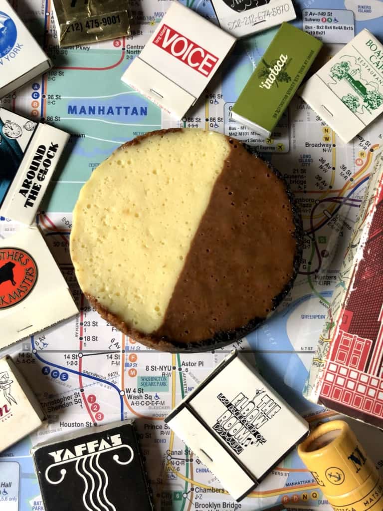 a NYC subway map with a mini black and white cookie-inspired cheesecake in the middle with matchbooks from some of nyc's iconic restaurants and bars lying around it and a red NYC notebook. The cheesecake is split right down the middle with a light white chocolate side and a dark milk chocolate side to emulate the iconic cookie.