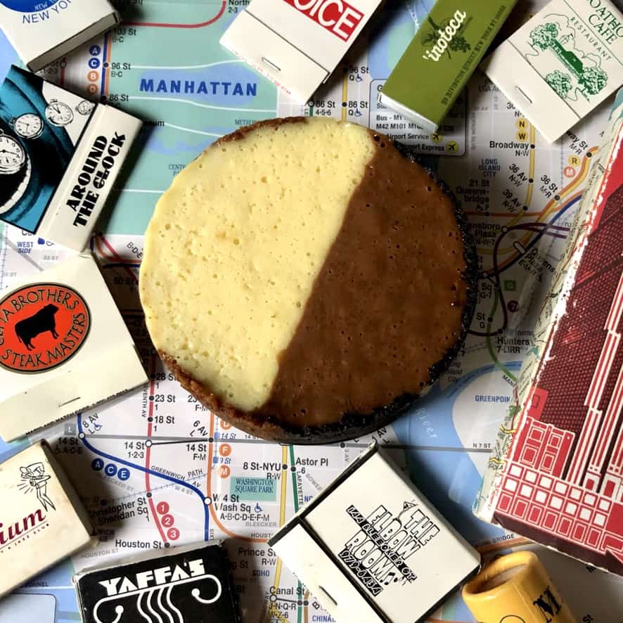 a NYC subway map with a mini black and white cookie-inspired cheesecake in the middle with matchbooks from some of nyc's iconic restaurants and bars lying around it and a red NYC notebook. The cheesecake is split right down the middle with a light white chocolate side and a dark milk chocolate side to emulate the iconic cookie.