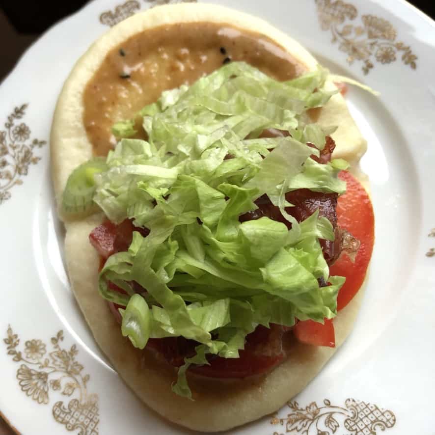 sesame soy mayo smeared over both sides of a bun and tomato slices on one half topped with crispy prosciutto and shredded lettuce