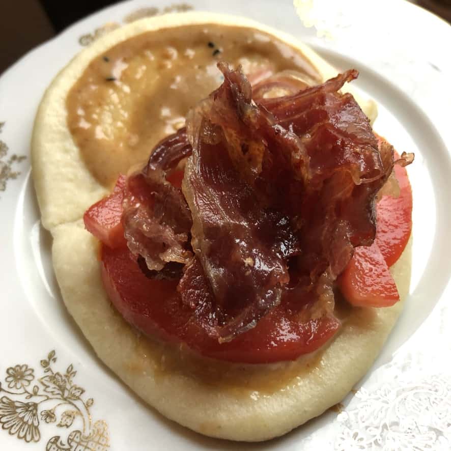 sesame soy mayo smeared over both sides of a bun and tomato slices on one half topped with crispy prosciutto