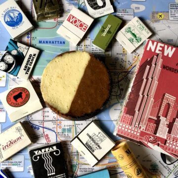 a NYC subway map with a mini black and white cookie-inspired cheesecake in the middle with matchbooks from some of nyc's iconic restaurants and bars lying around it and a red NYC notebook. The cheesecake is split right down the middle with a light white chocolate side and a dark milk chocolate side to emulate the iconic cookie.