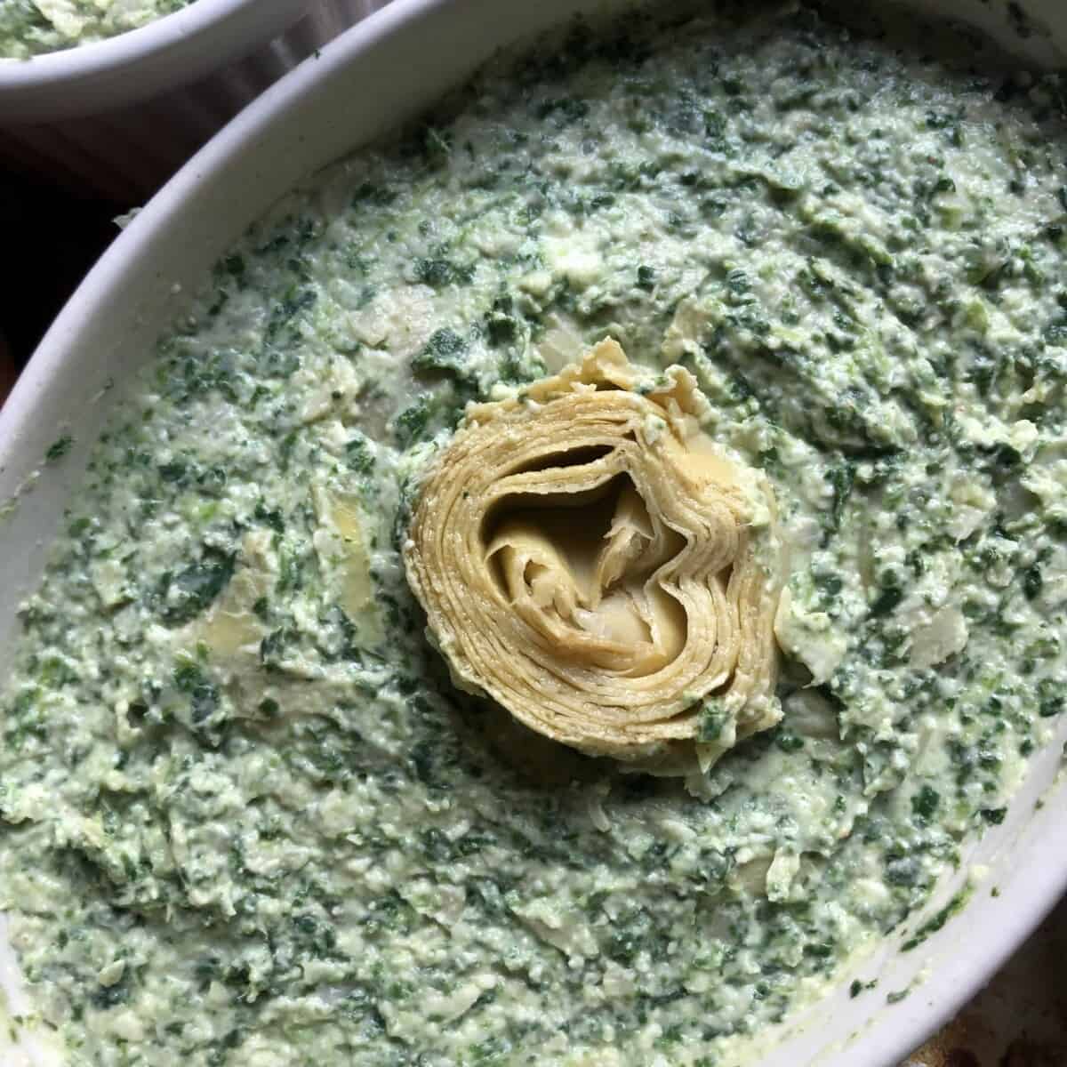 assembled unbaked oval ramekin filled with dip and an artichoke heart in the middle just barely sitting above the dip line