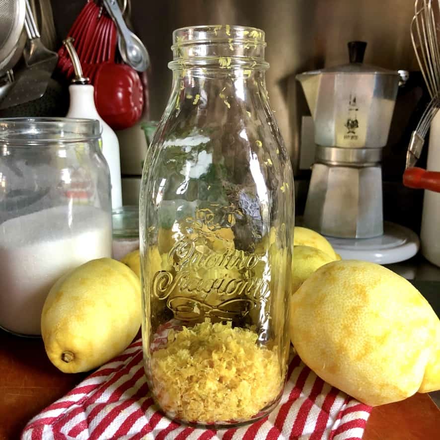 a large Italian canning jar (quattro stagione brand) filled with lemon zest and peels lying next to the zested lemons on a cutting board