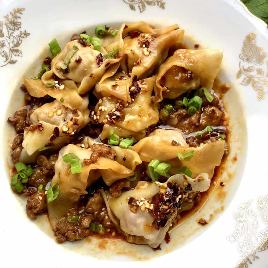 sichuan wontons resting in a bowl on top of sauce and topped with crispy fried pork and scallions all stirred together