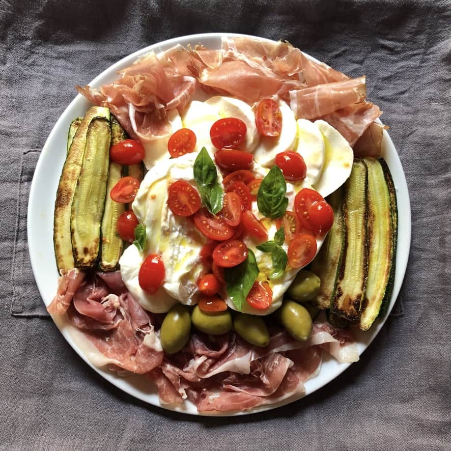 Antipasto platter with pan seared zucchini, sliced fresh grape Datterino tomatoes,, green olives, prociutto di Parma, Speck di Asiago, burrata, and buffalo mozzarella slices.