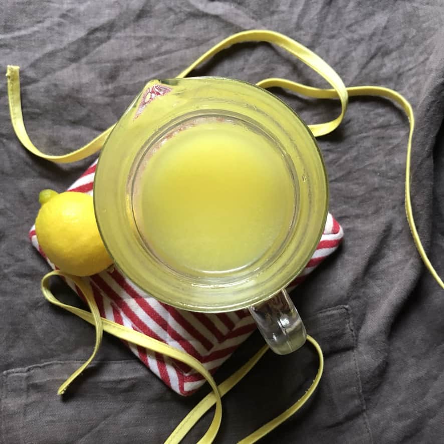 top down view of bright yellow limoncello in a glass pitcher with a fresh lemon next to it and yellow strings of the grey apron contrasting with the deep red and white striped pot holder it's sitting on top of (it looks creamy and buttery bright (almost flourescent yellow once the simple syrup is added)