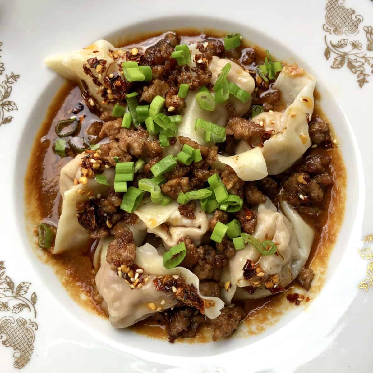 sichuan wontons resting in a bowl on top of sauce and topped with crispy fried pork and scallions without yet being mixed in together