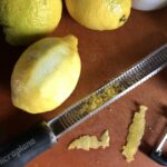 a Microplane zester lying next to some zested lemons with zest in the "canal" of the microplane and some peels lying next to the microplane (with zero pith on the peels)