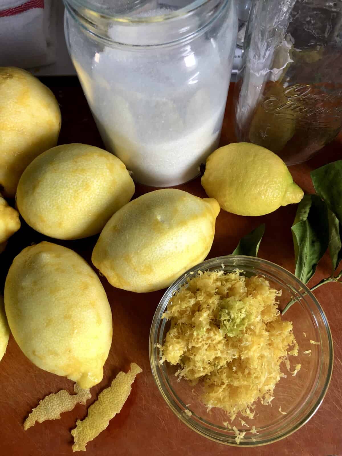 fully zested and peeled lemons lying on a cutting board with a sugar jar, alcohol, and a bowl full of zest