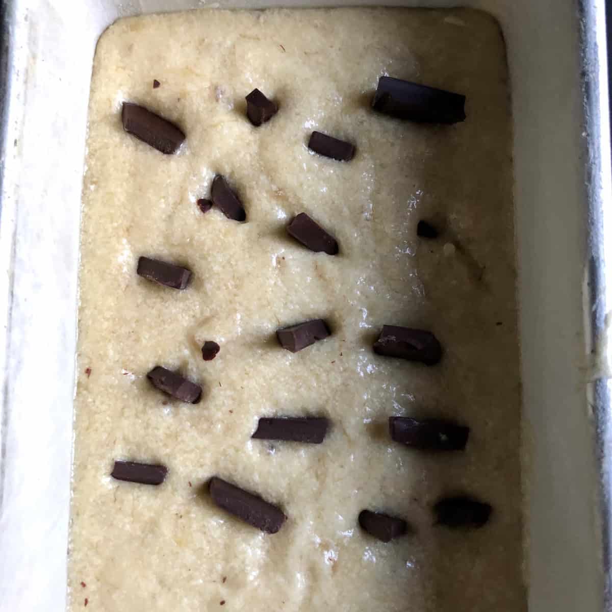 Chocolate chip banana bread batter in a loaf pan dotted with chunks of dark chocolate ready to bake, banana bread chocolate chips recipe from scratch.