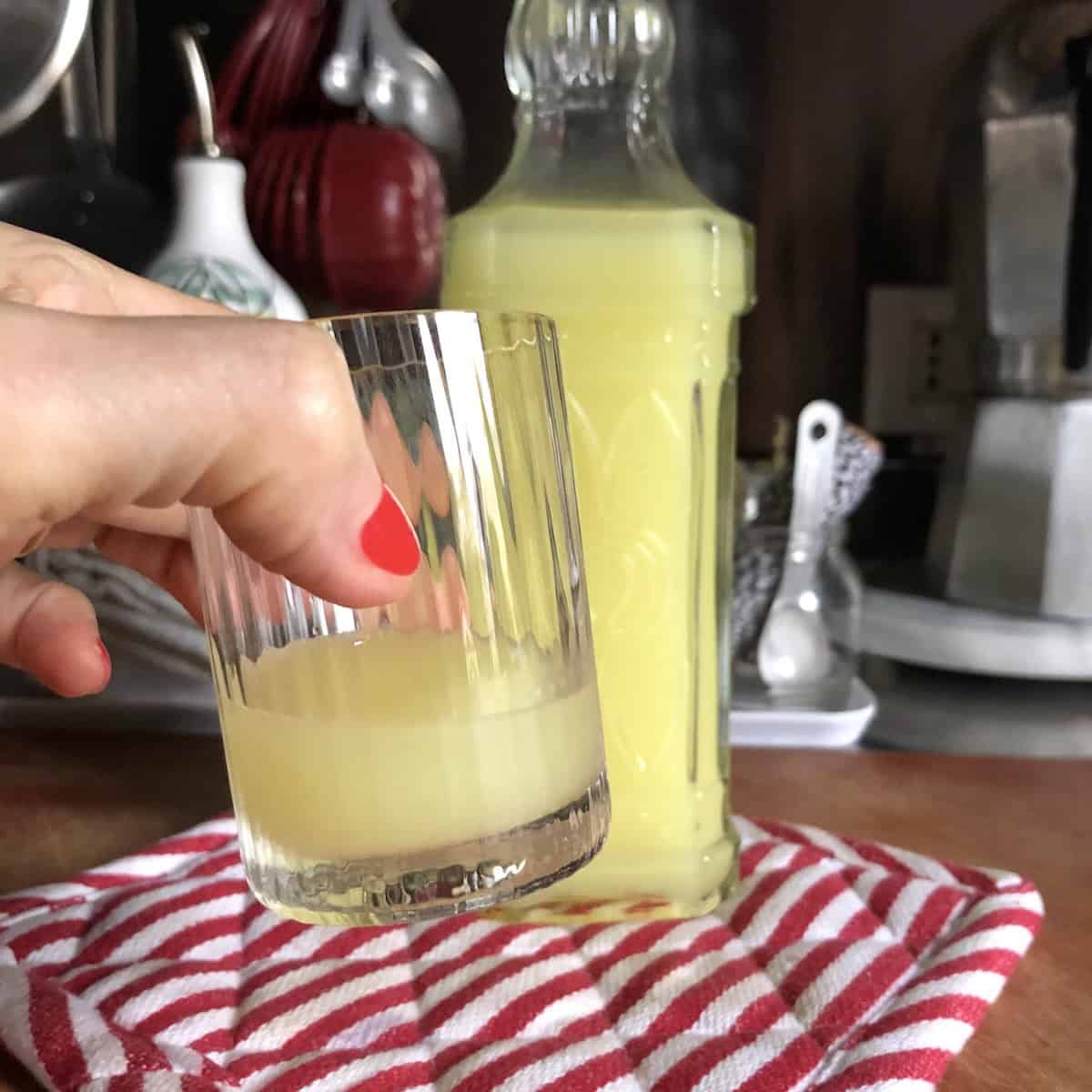 My hand holding a vintage Italian digestivo glass of my homemade limoncello in front of the limoncello bottle, served ice cold as a traditional Italian liqueur.