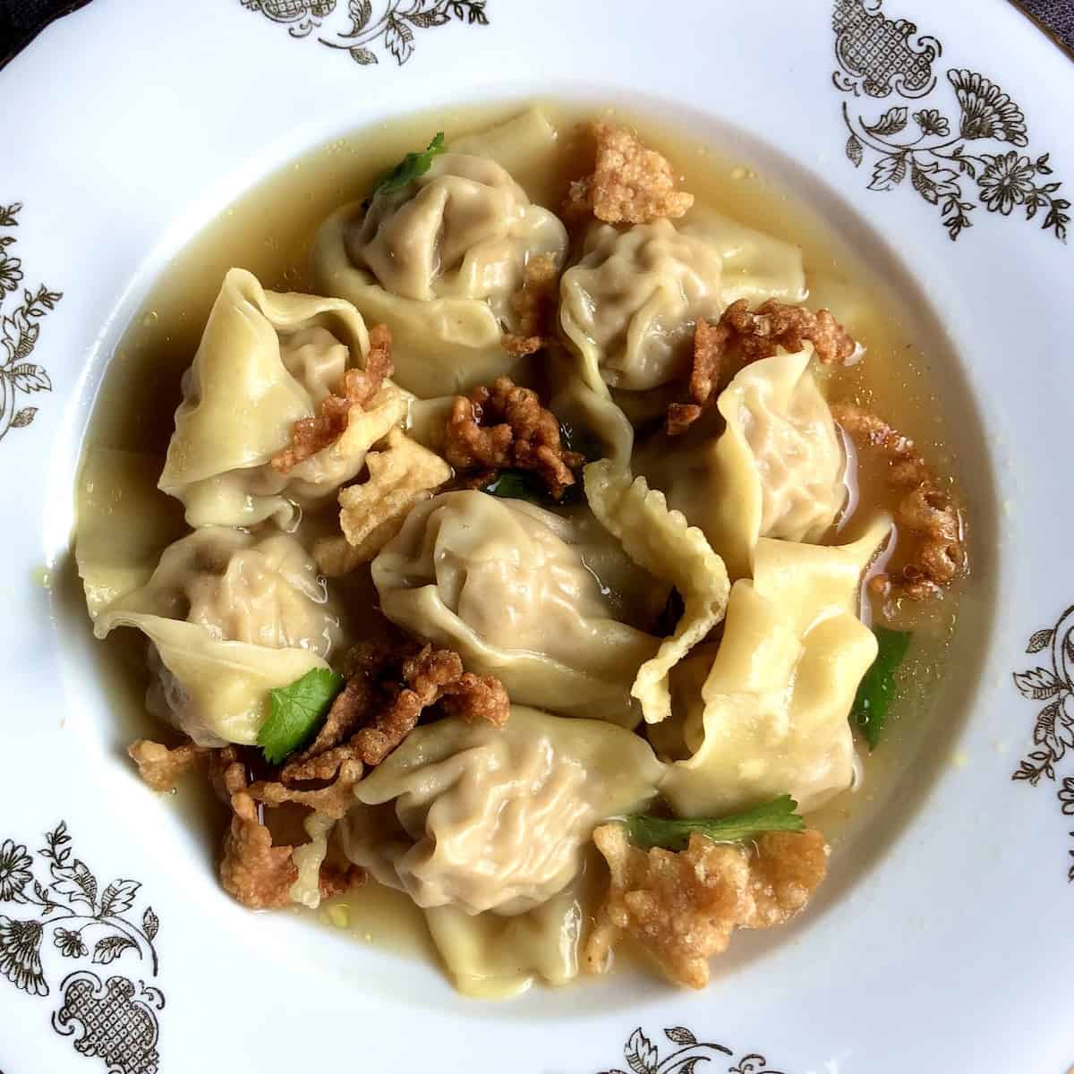 Traditional Chinese Wonton Soup in a bowl with homemade chicken broth glistening with bits of oil and fresh cilantro leaves and crispy fried homemade wonton strips.