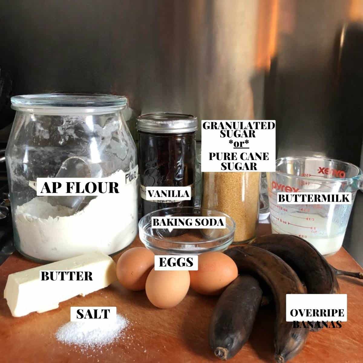 Chocolate chip banana bread ingredients on a cutting board with dark chocolate chunks, overly ripe bananas, flour, sugar, eggs, butter, banana bread with chocolate chips.