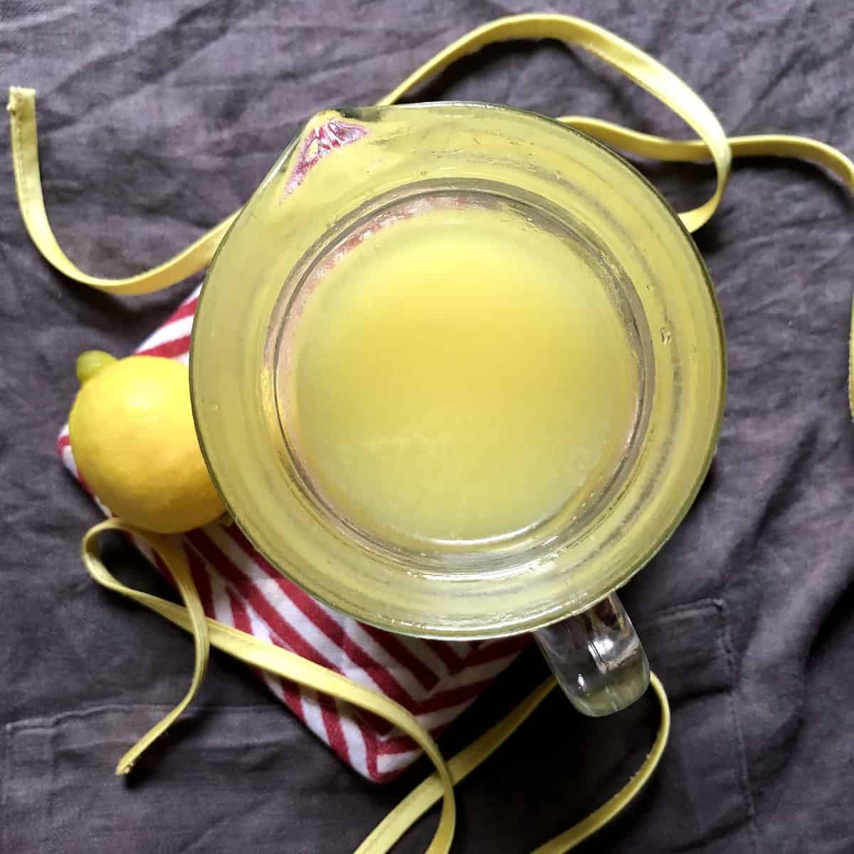 Top down view of bright creamy yellow limoncello in a glass pitcher with a fresh lemon, showing the fluorescent buttery color after adding the simple syrup, recipe for limoncello ready to bottle and cure.
