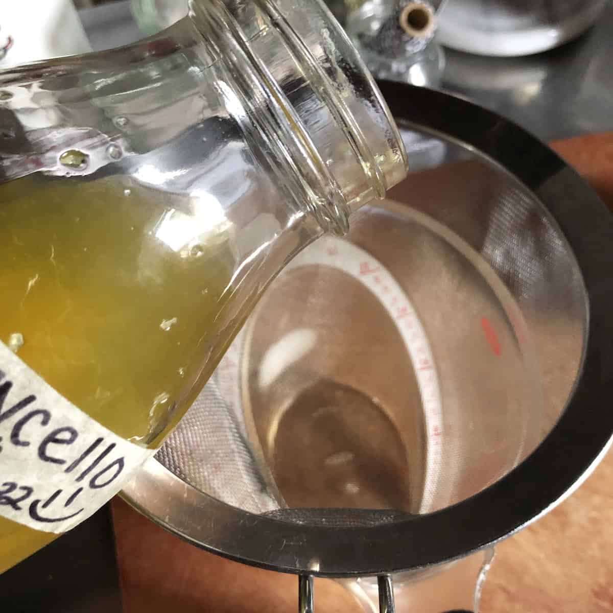 Straining the lemon zest and peels from the cured alcohol through a fine mesh strainer, limoncello recipes step showing how to make lemoncello clear and smooth before adding simple syrup.
