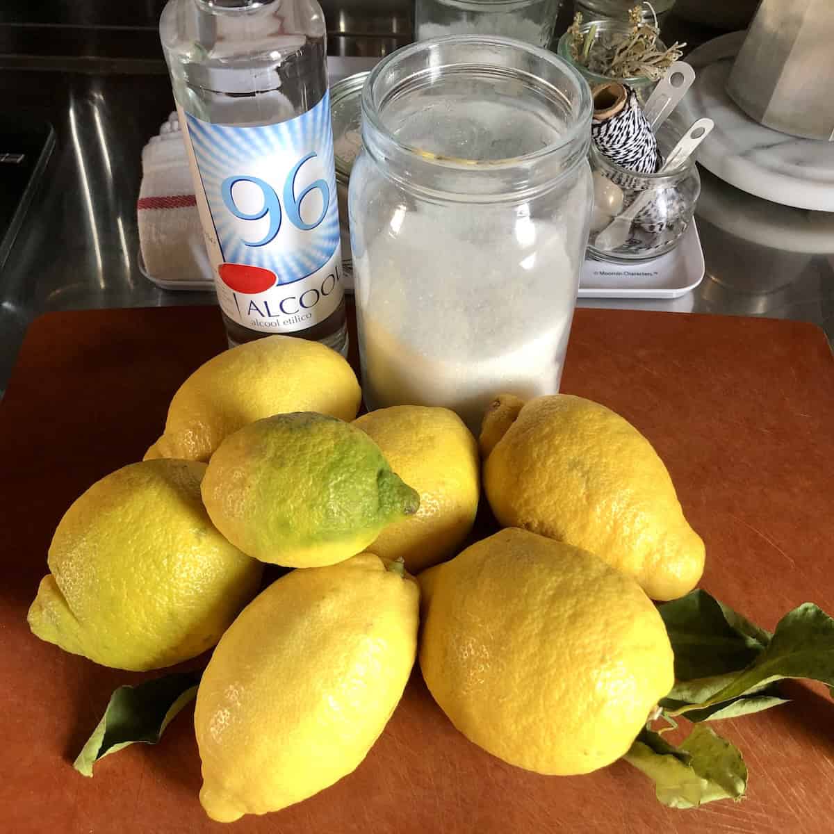 Homemade limoncello ingredients laid out on a cutting board including lemons, sugar, water, and pure grain alcohol, limoncello ingredients for an easy homemade limoncello recipe from scratch.

