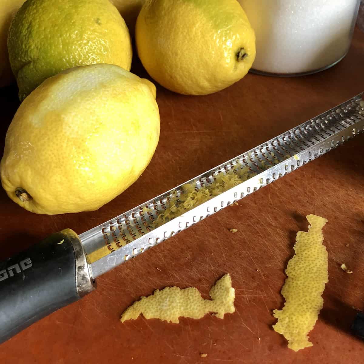 A Microplane zester lying next to zested lemons with fresh zest in the channel and lemon peels with zero pith, lemon zest for limoncello recipe using only the bright yellow outer peel.
