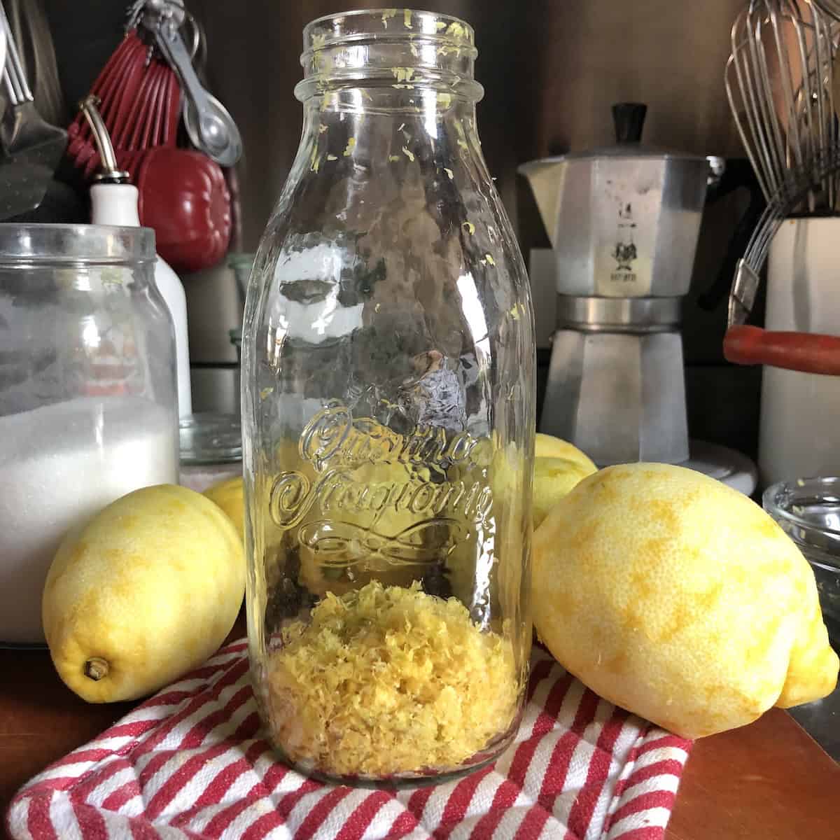 A large Italian Quattro Stagioni canning jar filled with lemon zest and peels lying next to zested lemons on a cutting board, limoncello ingredients ready for the alcohol infusion to make limoncello from scratch.
