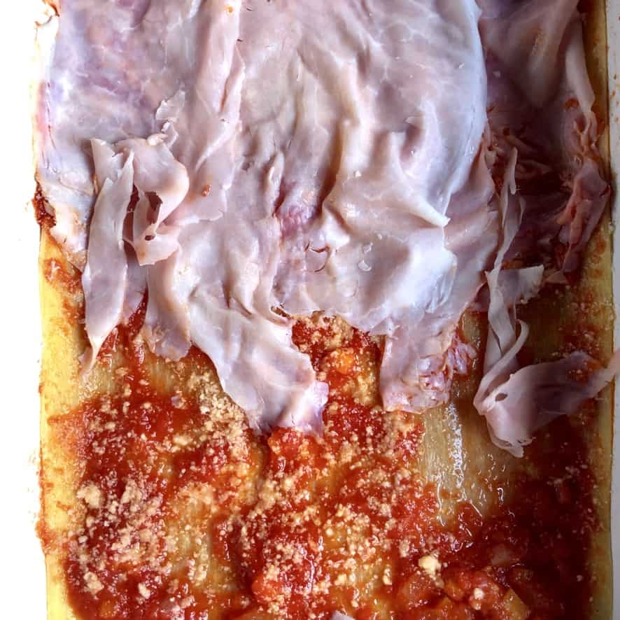 thin layers of Italian ham being layered on top of lasagna pasta sheets covered in tomato sauce