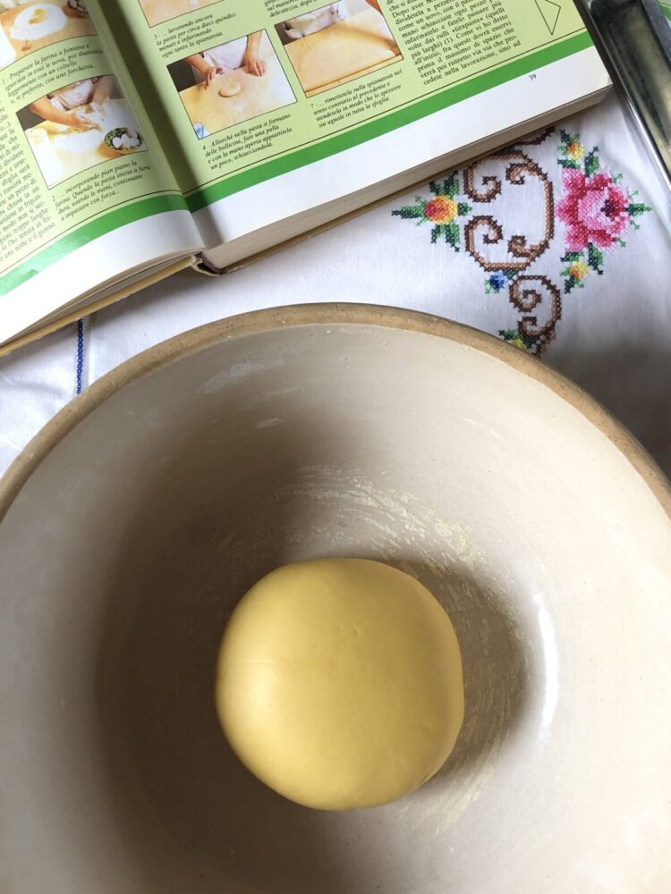 A well-hydrated, shiny, deep yellow-colored ball of homemade egg pasta in my great grandmother's bread bowl with a vintage Italian cookbook open to the tortellini recipe page