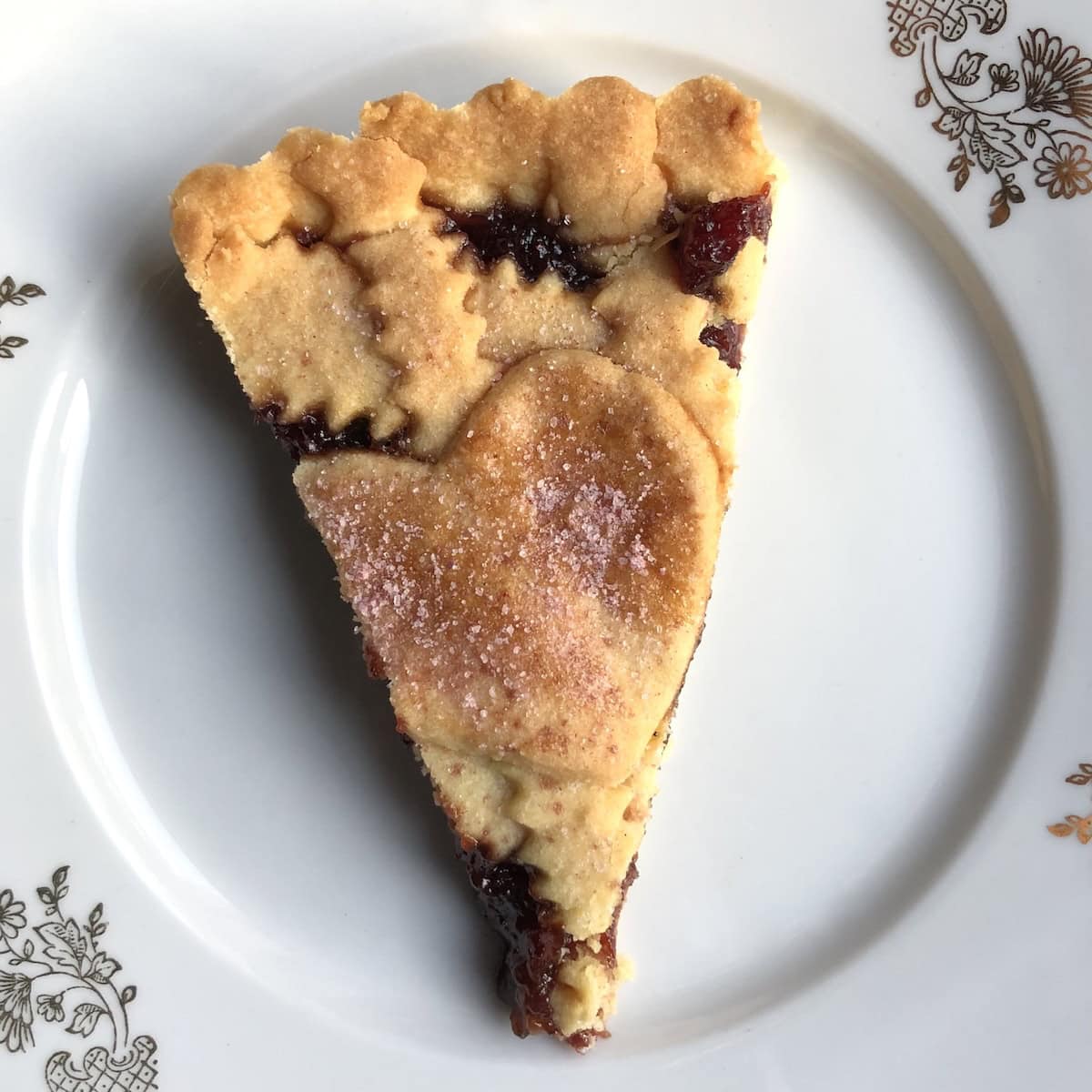 Slice of homemade cherry crostata (crostata alla ciliegia) with decorative heart cutouts on reginelle lattice top showing tender pasta frolla crust and ruby red cherry jam filling, authentic Italian jam tart recipe.